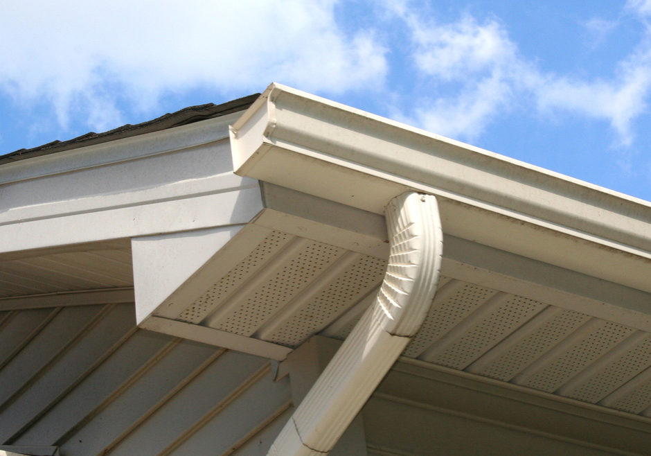Gutter and downspout of a house with a blue sky and white clouds in the background.